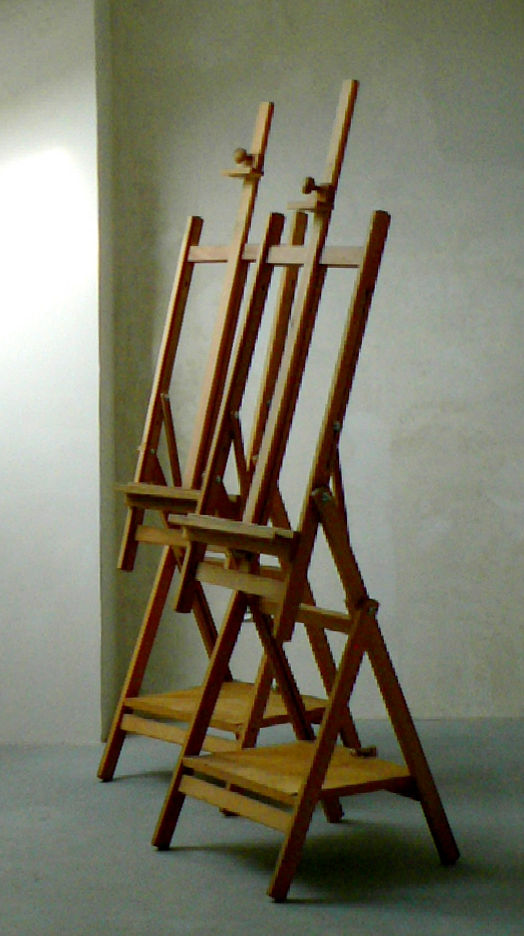 Fotografia przedstawia dwie sztalugi studyjne, drewniane w malowniczo oświetlonym pomieszczeniu.